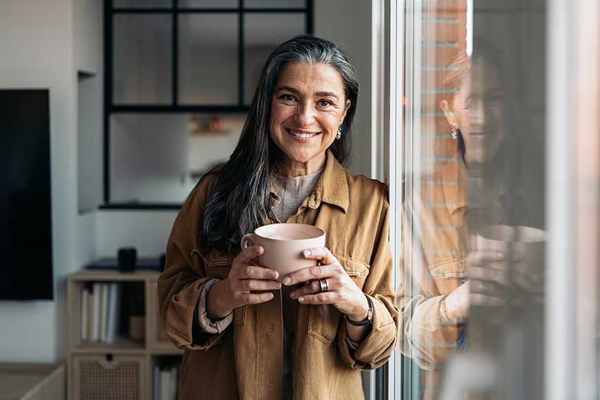 A person standing by a window holding a mug faces the camera